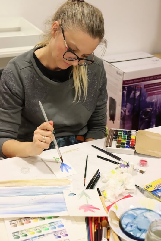 Skupiona uczestniczka maluje obraz. Widoczne delikatne kwiaty. Obok blok i różne kolorowe farby, a także dokończony pejzaż w ciepłych, letnich barwach.