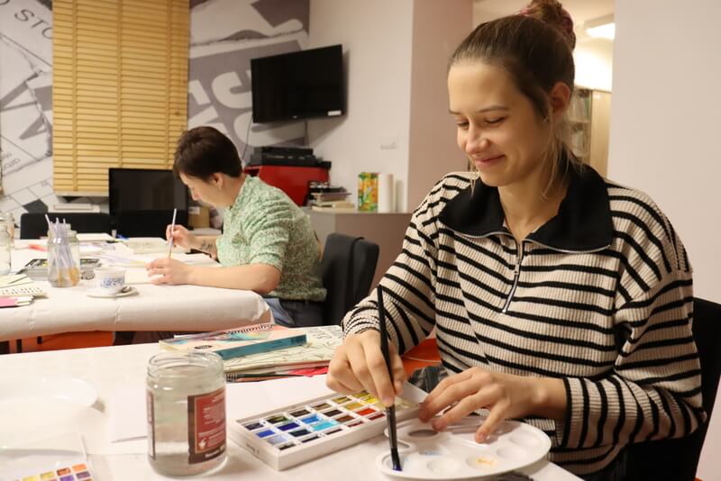 Zbliżenie na uśmiechniętą, młodą kobietę malującą kwiat. W tle widać inne uczestniczki zajęć. Przed malującą widoczne farby, papier, różowe zakładki DKK. Słoik z wodą.