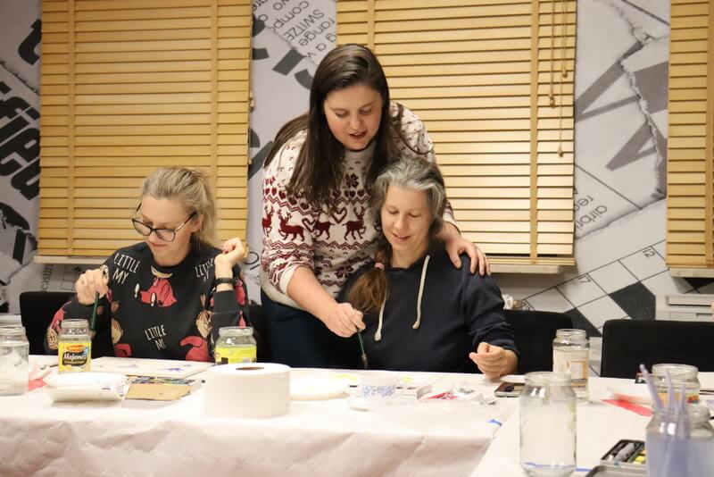 Prowadząca Melcher nachyla się nad skupioną uczestniczką i daje wskazówki, jak poprowadzić pędzel po pracy. Obok siedzi inna młoda kobieta i maluje swój obraz. Przed dziewczynami kolorowe farby, pędzle, słoiki z wodą.