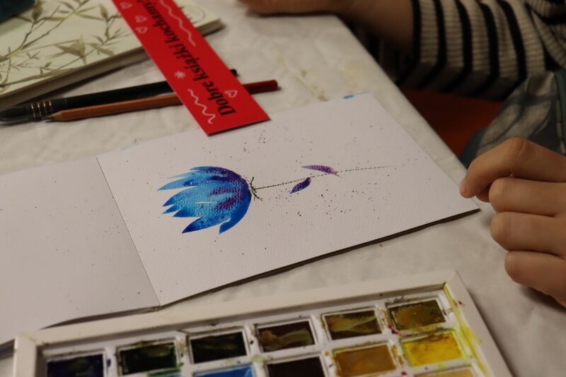 Zbliżenie na piękny obrazek przedstawiający niebieski kwiat. Obok pędzel, farby. Różowa zakładka DKK.