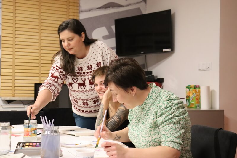 Ujęcie na dwie uśmiechnięte uczestniczki maczające pędzle w wodzie. Pomiędzy nimi prowadząca pomagająca w pracy. Przed nią leży rozpoczęty obraz. W tle podparta inna uczestniczka malująca soją pracę. Przed nimi farby, pędzle, słoiki z wodą.
