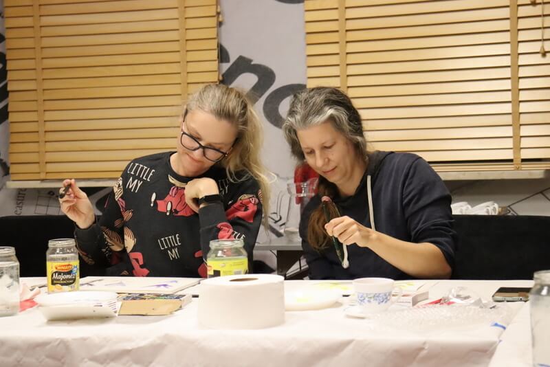 Czytelnia Działu Regionalnego. Dwie skupione na malowaniu młode kobiety siedzą przy stole nakrytym białym obrusem papierowym. Przed nimi leżą kolorowe farby, pędzle, słoiki z wodą. W tle drewniane żaluzje w oknach.