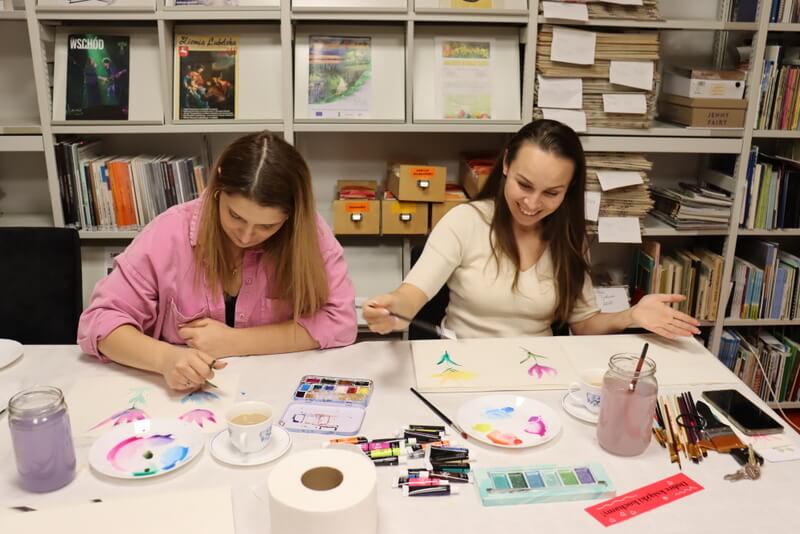 Czytelnia Działu Regionalnego. Dwie uśmiechnięte młode kobiety siedzą i malują obrazki przy stole nakrytym białym obrusem papierowym. Przed nimi leżą kolorowe farby, pędzle, słoiki z wodą. W tle regały z prasą regionalną i książkami.