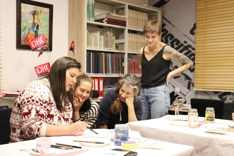 Czytelnia Działu Regionalnego. Dwie uśmiechnięte młode kobiety siedzą przy prowadzącej warsztaty K. Melcher, która pokazuje instruktaż do pracy z akwarelą. Siedzą przy stole nakrytym białym obrusem papierowym. Przed nimi leżą kolorowe farby, pędzle, słoiki z wodą. Jedna z kobiet stoi i zerka z góry na malowaną pracę. W tle na ścianie wisi kolorowy obraz, a w oknie wiszą drewniane żaluzje. Regały z książkami regionalnymi.