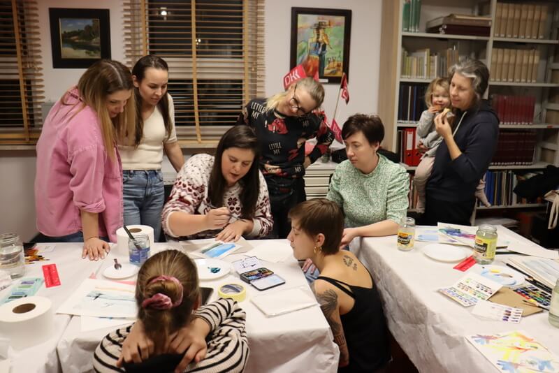 Czytelnia Działu Regionalnego. Stoły przykryte białymi obrusami papierowymi. Na stołach kolorowe farby, kartki papieru i rysunki. Kobiety zgromadzone dookoła prowadzącej warsztaty patrzą i słuchają, jak K. Melcher rysuje instruktażowy obraz. Młode kobiety zgromadzone wokół prowadzącej zainteresowane słuchają. Jedna ze stojących kobiet trzyma na rękach dziecko. W tle różowe chorągiewki z logo Dyskusyjnego Klubu Książki.