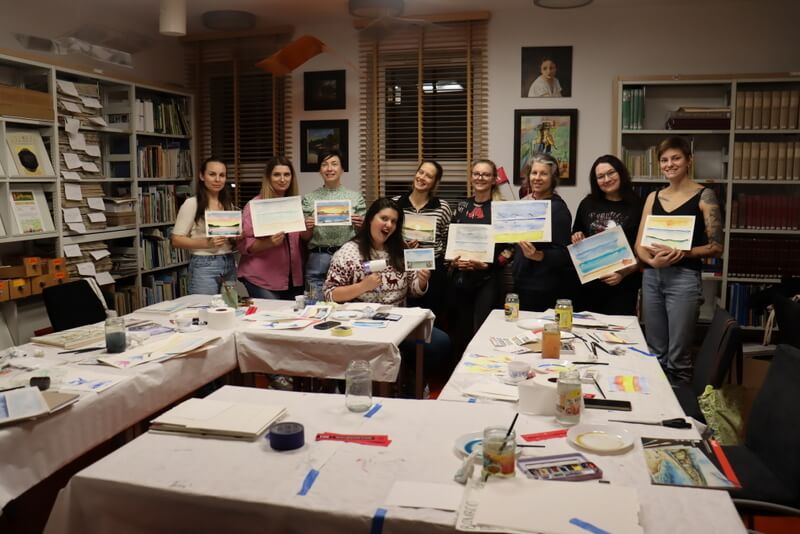 Czytelnia Działu Regionalnego. Zdjęcie grupowe, na którym pozują uśmiechnięte młode kobiety trzymające swoje obrazki malowane akwarelą. Dziewięć kobiet i w centrum prowadząca warsztaty K. Melcher, pozująca z suszarką do włosów – używaną do osuszania nadmiaru wody z papieru do akwareli. Na pierwszym planie stoły, na których leżą narzędzia do malowania: kolorowe farby, pędzle, słoiki z wodą, palety do mieszania kolorów.