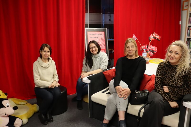Oddział dla Dzieci. Przy białym stole, na kanapie a la fortepian, siedzą 4 uśmiechnięte kobiety. Pozują do zdjęcia. Dwie z nich siedzą na pufach, po lewej stronie widoczne pluszowe siedziska - żółte misie. W tle czerwona kotara i różowy plakat z logo DKK, na białym stole z tyłu chorągiewki z tym samym logo.
