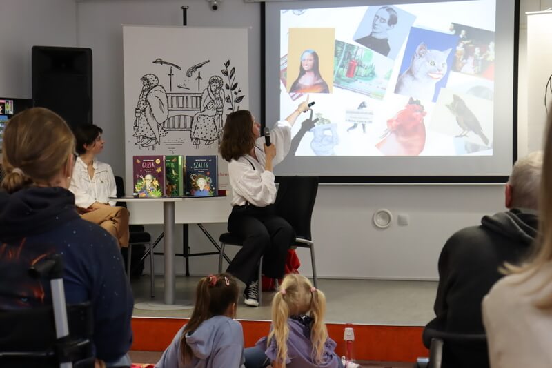 Na pierwszym planie siedzi publiczność wsłuchana w prezentację. Na scenie siedzą Joanna Rusinek i Urszula Panasiuk. Joanna Rusinek trzyma mikrofon i wskazuje dłonią na slajd wyświetlony na ekranie, przedstawiający kolażowe ilustracje. Na stoliku stoją trzy kolorowe książki.