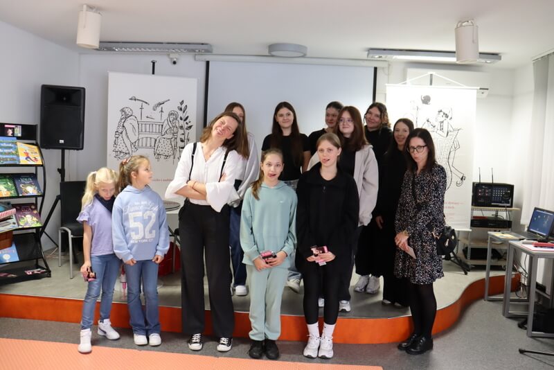 Na zdjęciu widać grupę uczestników spotkania, stojących na scenie w bibliotece. W centrum znajduje się Joanna Rusinek. Wszyscy pozują do pamiątkowego zdjęcia robiąc głupie miny.
