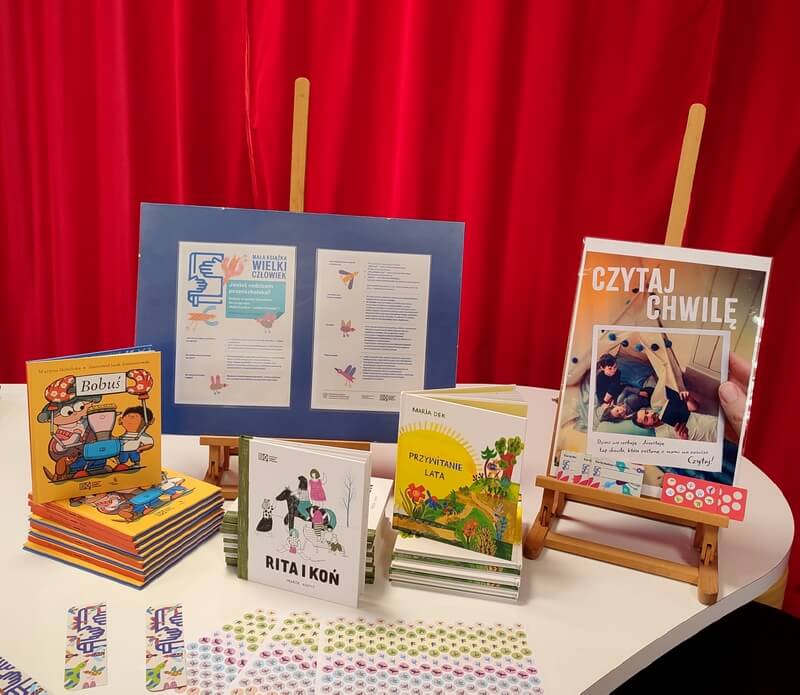 Na zdjęciu widnieje plakat z&nbsp;logo projektu Mała Książka – Wielki Człowiek. Przed plakatem wyeksponowane są wyprawki czytelnicze, które przedszkolaki otrzymają podczas tej edycji. Tło w&nbsp;kolorze czerwonym.