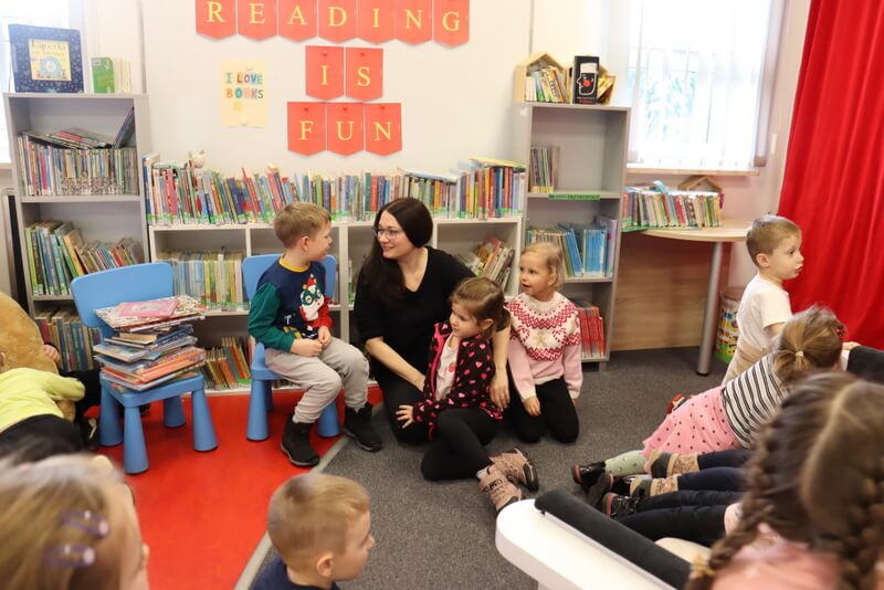 Oddział dla Dzieci. Poniżej białej magnetycznej tablicy, na której wisi żółty napis na czerwonym tle „Reading is fun” siedzi grupka dzieci i prowadząca spotkanie. Uśmiechają się do siebie i rozmawiają. Chłopiec siedzi na niebieskim krzesełku i uśmiecha się do prowadzącej, opowiada coś wesoło. Obok po prawej stronie dwie dziewczynki przytulają się do prowadzącej. W tle za nimi regały z kolorowymi książeczkami.