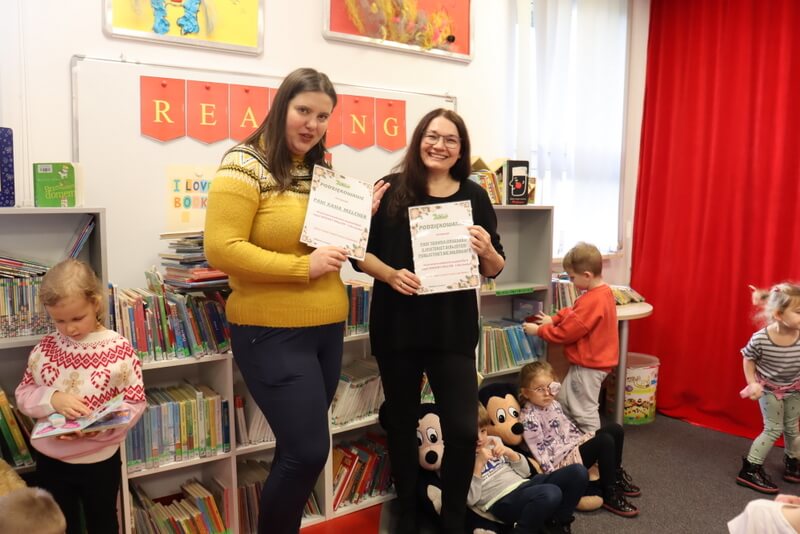 Oddział dla Dzieci. Dwie kobiety, prowadząca spotkanie i autorka prac prezentowanych na wystawie uśmiechnięte pozują do zdjęcia trzymając podziękowanie od przedszkolaków. W tle za nimi wisi na ścianie biała tablica magnetyczna z żółtym napisem na czerwonym tle „Reading is fun”. Po bokach stoją dzieci z książeczkami, regały z kolorowymi książkami dla dzieci. Po prawej stronie fragment czerwonej kotary.