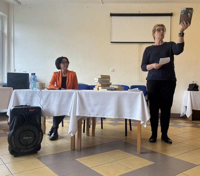 Bibliotekarka stoi przy stole z książką w ręce, za stołem z książkami siedzi druga bibliotekarka biorąca udział w spotkaniu.