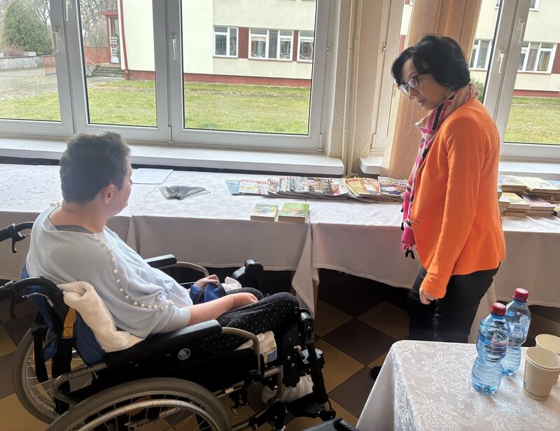 Przy stole z książkami siedzi na wózku uczestniczka spotkania, obok stoi bibliotekarka.