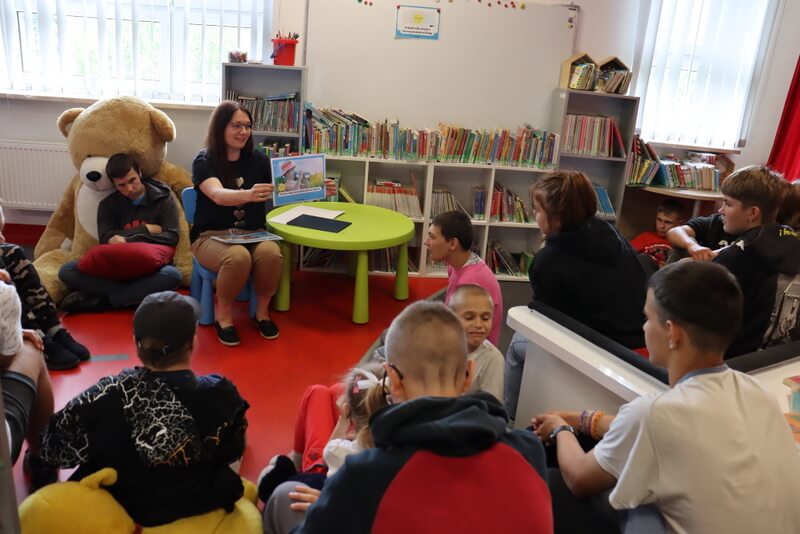 Oddział dla Dzieci. Grupa młodzieży patrzy na prowadzącą, która siedzi na małym, plastikowym niebieskim krzesełku i pokazuje obrazek z sytuacją wakacyjną. W tle na ścianie wisi biała tablic magnetyczna, na której przypięty jest inny rysunek z sytuacją wakacyjną. Obok prowadzącej stoi okrągły, zielony stolik plastikowy. Nieco z tyłu za prowadzącą, po lewej stronie na ogromnym beżowym misiu siedzi chłopak i uśmiecha się do obiektywu.