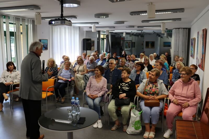 Publiczność siedząca na krzesełkach. Przed nimi stoi mężczyzna z mikrofonem.