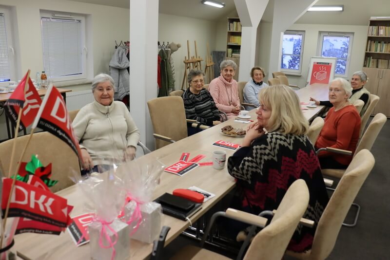 Duża sala, przy stole siedzi grupa kobiet, w tle okna a przy ścianie stoi regał z książkami. Na stole leżą materiały promocyjne, stoi talerz z ciastkami i wazon z chorągiewkami z logo DKK.