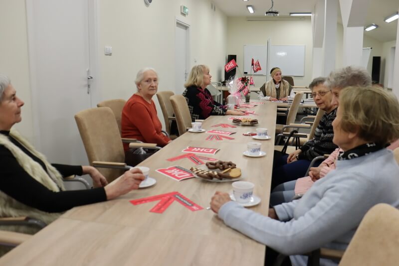 Przy stole siedzi grupa kobiet, na stole leżą materiały promocyjne, stoją filiżanki, talerz z ciastkami i wazon z chorągiewkami z logo DKK.