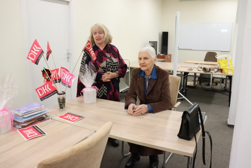 Duża sala, w której znajdują się dwie kobiety. Jedna siedzi a druga stoi i coś mówi. Na stole leżą książki i stoi flakon z chorągiewkami z logo DKK.