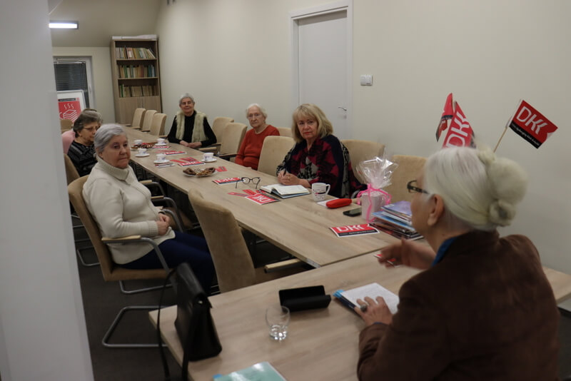 Grupa kobiet siedzi przy stole, jedna z nich mówi coś do reszty. Na stole leżą materiał promocyjne, książki, stoi talerz z ciastkami, filiżanki i wazon z chorągiewkami z logo DKK.