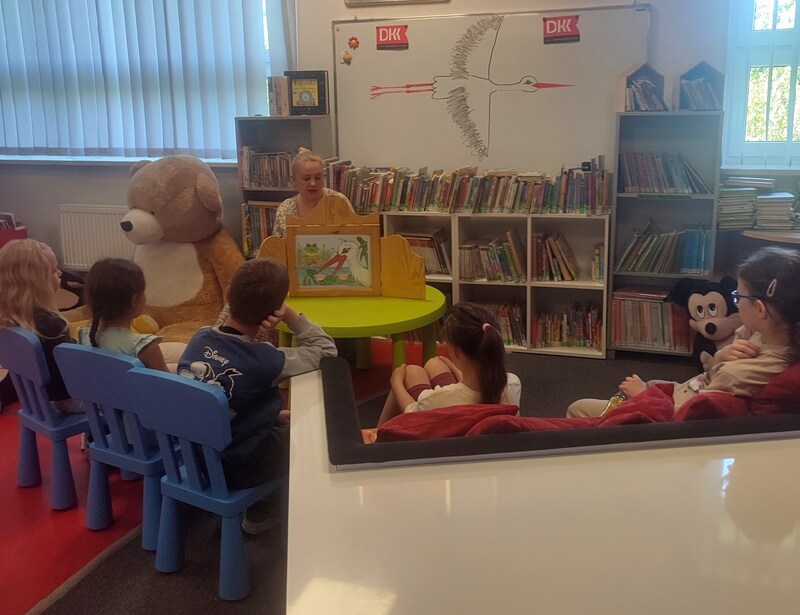 Grupa dzieci skierowana twarzami do bibliotekarki, która przedstawia dzieciom opowieść w teatrzyku kamishibai. Tło stanowi wystrój biblioteki w postaci tablicy na której widnieje wizerunek Bociana a także regały z książkami.