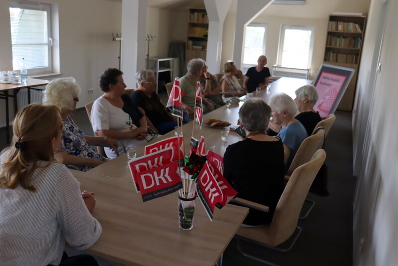 Pokój naukowy. Przy długim stole siedzi jedenaście kobiet. Kobieta na pierwszym planie siedzi tyłem. Obok, na stole, chorągiewki z logo DKK.
