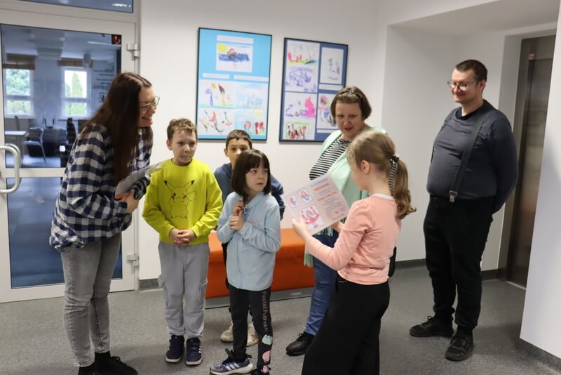 Hol Galerii na Piętrze. Prowadząca spotkanie wręcza podziękowanie dla Artystki – Emilii. Obok stoją uśmiechnięci rodzice dziewczynki. W&nbsp;tle po lewej stronie zamknięte, przeszklone  drzwi do Czytelni Internetowej. Po prawej stronie wejście do windy. Na ścianach powieszone równo antyramy z&nbsp;kolorowymi rysunkami. Pod ścianą stoją pomarańczowe kwadratowe pufy.