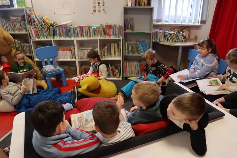 Widok na kącik czytelniczy w Oddziale dla Dzieci. Grupa przedszkolaków ogląda książki żywo gestykulując i pokazując sobie wzajemnie ilustracje.