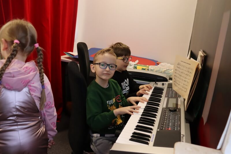 Dwóch chłopców siedzi przy keyboardzie i gra na instrumencie. Pozują do zdjęcia. Keyboard stoi za czerwoną kotarą, w tle widoczne kolorowe kartony ułożone na stoliku za chłopcami.