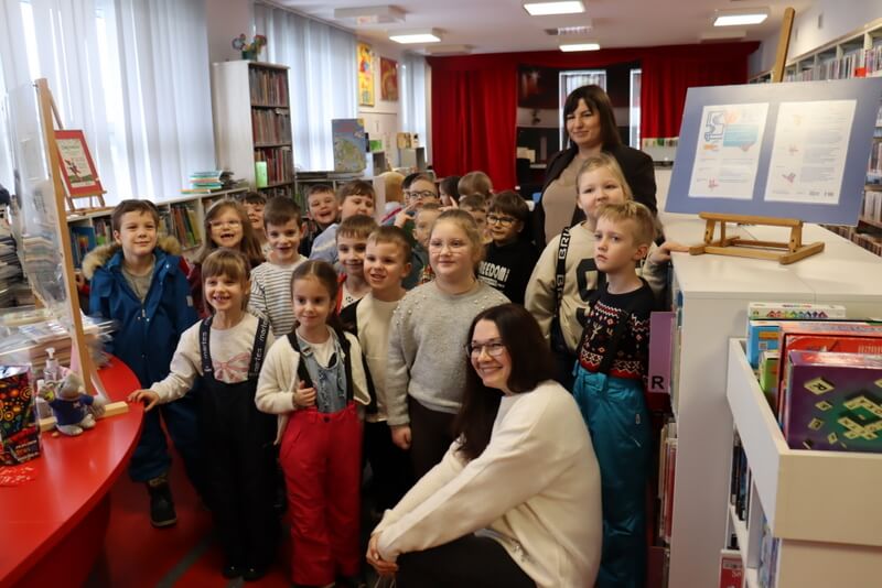 Oddział dla Dzieci, widok na wypożyczalnię. Grupa dzieci stoi wzdłuż pomieszczenia i pozuje do zdjęcia wraz z paniami: wychowawczynią i prowadzącą, która kuca przy dzieciach. Uśmiechają się do zdjęcia.