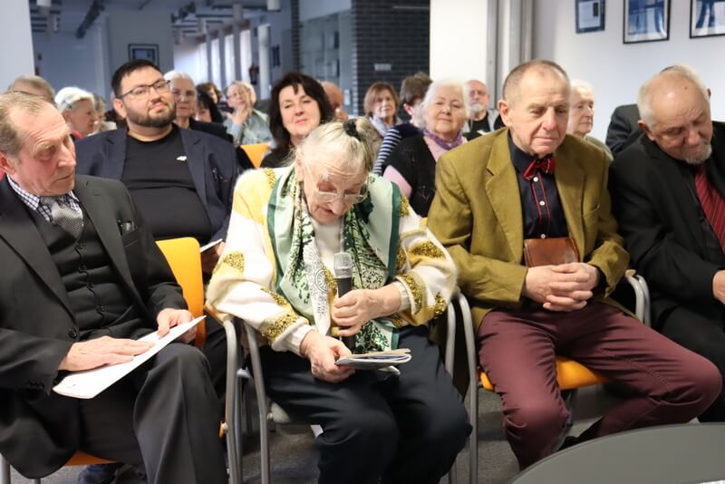 Grupa ludzi siedzących na krzesłach. Kobieta czyta do mikrofonu.