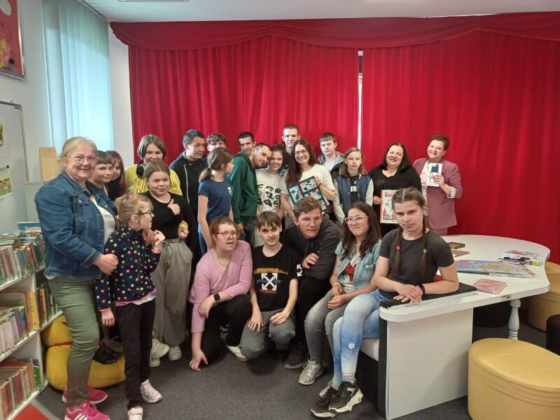 Zdjęcie grupowe na tle czerwonej kotary w&nbsp;Oddziale dla Dzieci. Wśród roześmianej młodzieży stoją nauczyciele/wychowawcy oraz prowadząca spotkanie J. Orzeszko. Część osób siedzi po prawej stronie na kanapie w&nbsp;kształcie fortepianu.