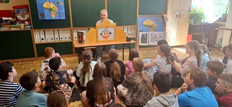 Na pierwszym planie grupa dzieci skierowana jest w kierunku prowadzącej, która przedstawia im ilustrowaną opowieść w teatrzyku kamishibai. W tle widnieją plansze edukacyjne.