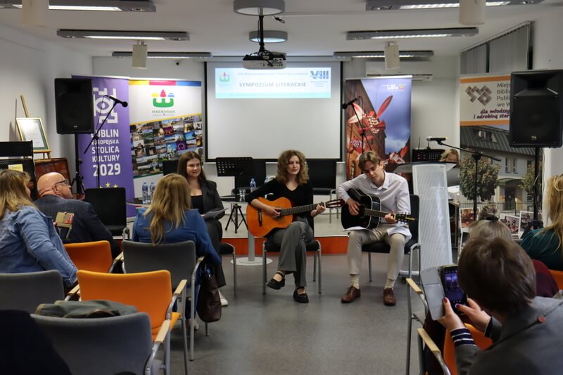 Dwie dziewczyny i jeden chłopak siedzący na krzesłach przed publicznością. Dziewczyna po środku i chłopak po jej lewej stronie grają na gitarach