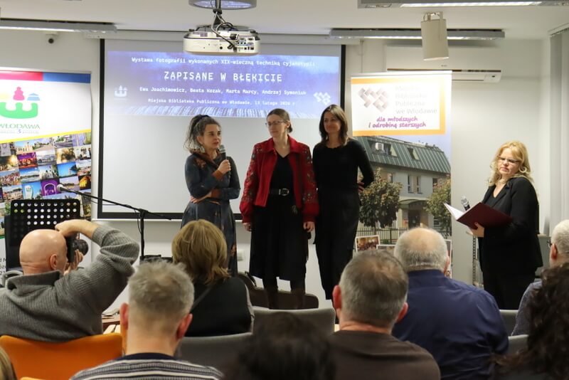 Trzy kobiety stojące na scenie. Poniżej stoi jeszcze jedna kobieta. Przed nimi siedzą ludzie na krzesłach.